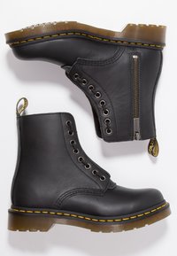 Botas de tobillo de cuero negro con costuras amarillas, parte delantera con cordones, cremallera lateral en una bota y suelas de goma marrón translúcidas.