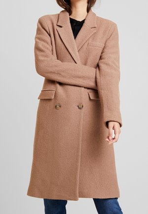 Kvinde iført en lang, dobbeltknappet camel-farvet uldfrakke over mørke bukser, med armene krydsede foran en grå baggrund.