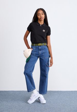 Polo shirt nero con colletto zip, jeans cargo a gamba larga blu, cintura verde e sneaker bianche. In una mano tiene un cappellino crema e verde.