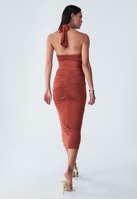Rust-colored halter neck dress with a fitted silhouette, textured fabric, and ruching details along the back, ending below the knee.