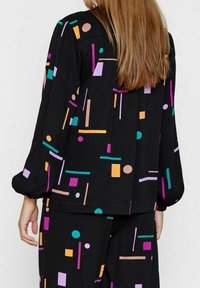 Černá látková košile s barevnými geometrickými vzory v purpurové, růžové, oranžové, zelené a modré; má dlouhé rukávy a volný střih.