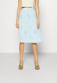 Light blue, pleated skirt with floral print in yellow and green hues. Made from a textured fabric, falls to mid-calf, paired with green sandals.