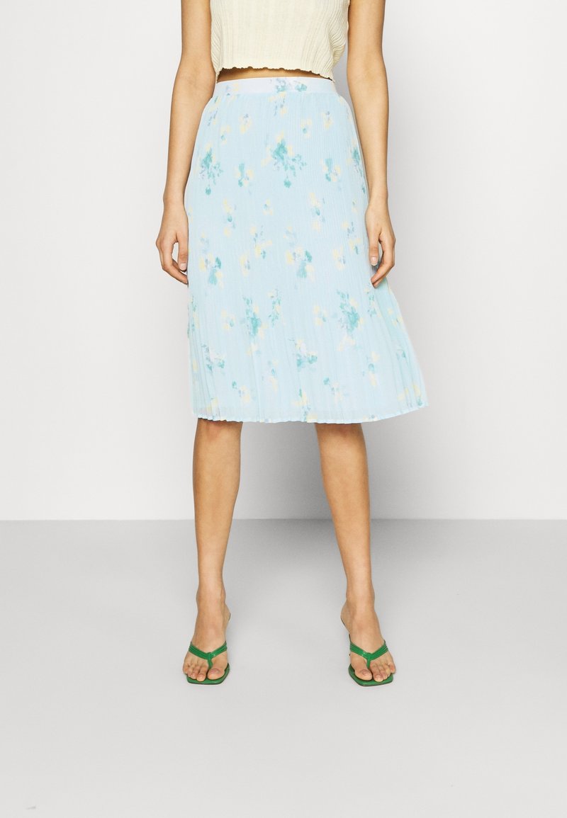 Light blue, pleated skirt with floral print in yellow and green hues. Made from a textured fabric, falls to mid-calf, paired with green sandals.