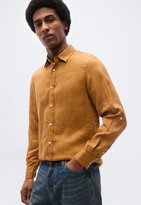 Άνδρας που φοράει πουκάμισο μουσταρδί με μακριά μανίκια και μπλε τζιν, με το ένα χέρι στην τσέπη, στέκεται μπροστά σε απλό ανοιχτό φόντο.