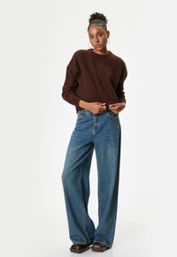 Maglione marrone lavorato a maglia con scollatura rotonda, abbinato a jeans blu a gamba larga. Scarpe con piattaforma marroni completano l'outfit.