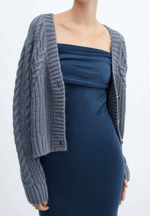 Personne portant une robe bleu marine sans bretelles avec un cardigan gris en maille torsadée ample posé sur les épaules, bras le long du corps.