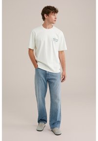 Lichtcreme t-shirt met een grafisch ontwerp "EXPLORE" op de borst, gecombineerd met lichtblauwe loszittende jeans en grijze instapschoenen.