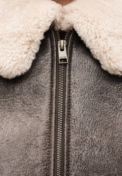 Καφέ δερμάτινο μπουφάν με γιακά από κρεμ χρώματος shearling, κλείσιμο με φερμουάρ με ασημί τράβηγμα "FRAME" και υφασμένη επιφάνεια.