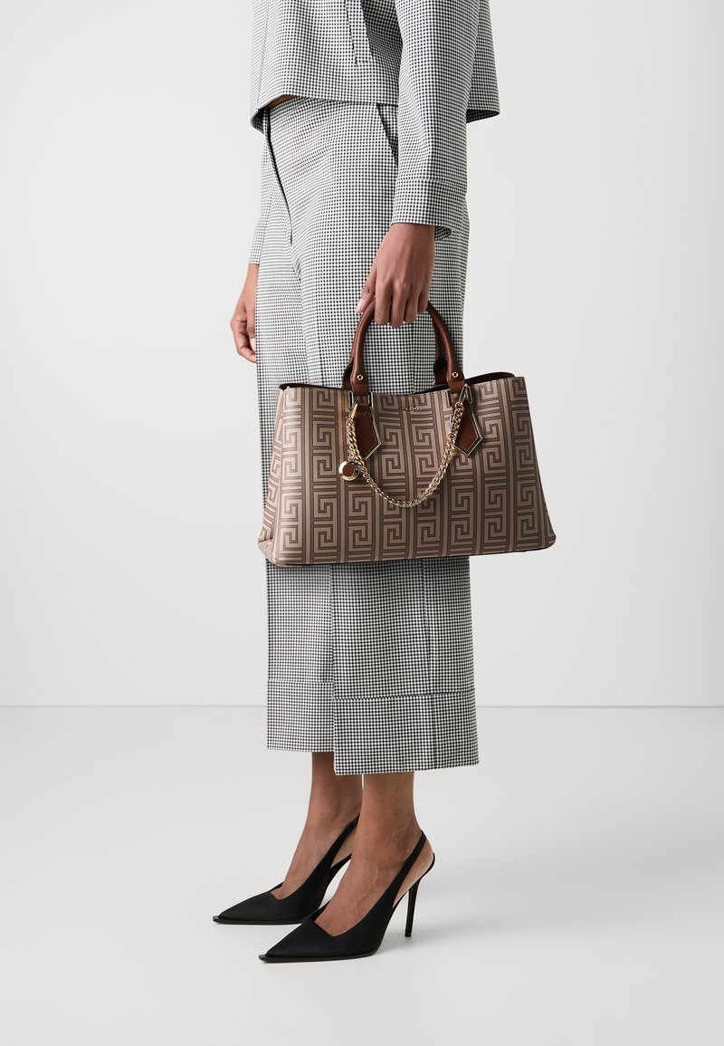 Sac à main marron à motif avec une bandoulière en chaîne et des poignées en cuir, présentant un design géométrique et une base carrée, tenu par une personne en tenue à carreaux.