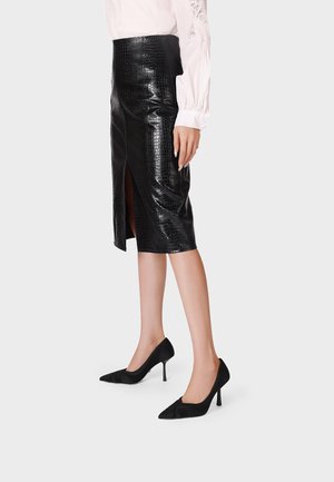 Donna che indossa una gonna midi in pelle nera testurizzata con spacco frontale e scarpe nere con tacco alto e punta affilata, in piedi su uno sfondo uniforme.
