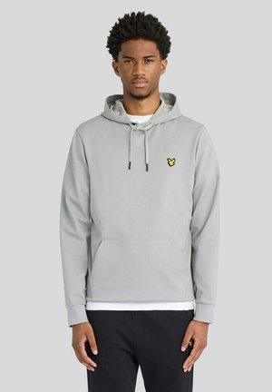 Junger Mann mit kurzen lockigen Haaren, trägt einen hellgrauen Hoodie mit gelbem Logo auf der Brust und schwarzen Hosen, steht vor einem einfachen hellen Hintergrund.