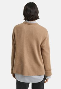 Pull léger marron avec un ourlet et des poignets côtelés, fabriqué dans un matériau doux, offrant une coupe décontractée sur une chemise à rayures bleues et blanches.
