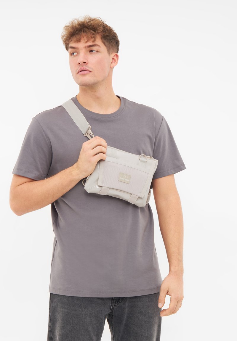 Borsa a tracolla grigio chiaro realizzata in materiale resistente, dotata di tasca frontale e tracolla regolabile. Texture liscia con design minimalista.