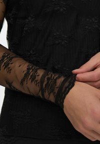 Čierny čipkový top s textúrovanou tkaninou, zložitými kvetinovými vzormi a priesvitnými dlhými rukávmi s jemnými vykrojenými manžetami.