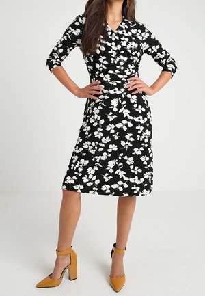 Frau trägt ein schwarzes knielanges Kleid mit weißem Blumenmuster und senfgelben High Heels mit Knöchelriemen, steht mit den Händen in der Hüfte.
