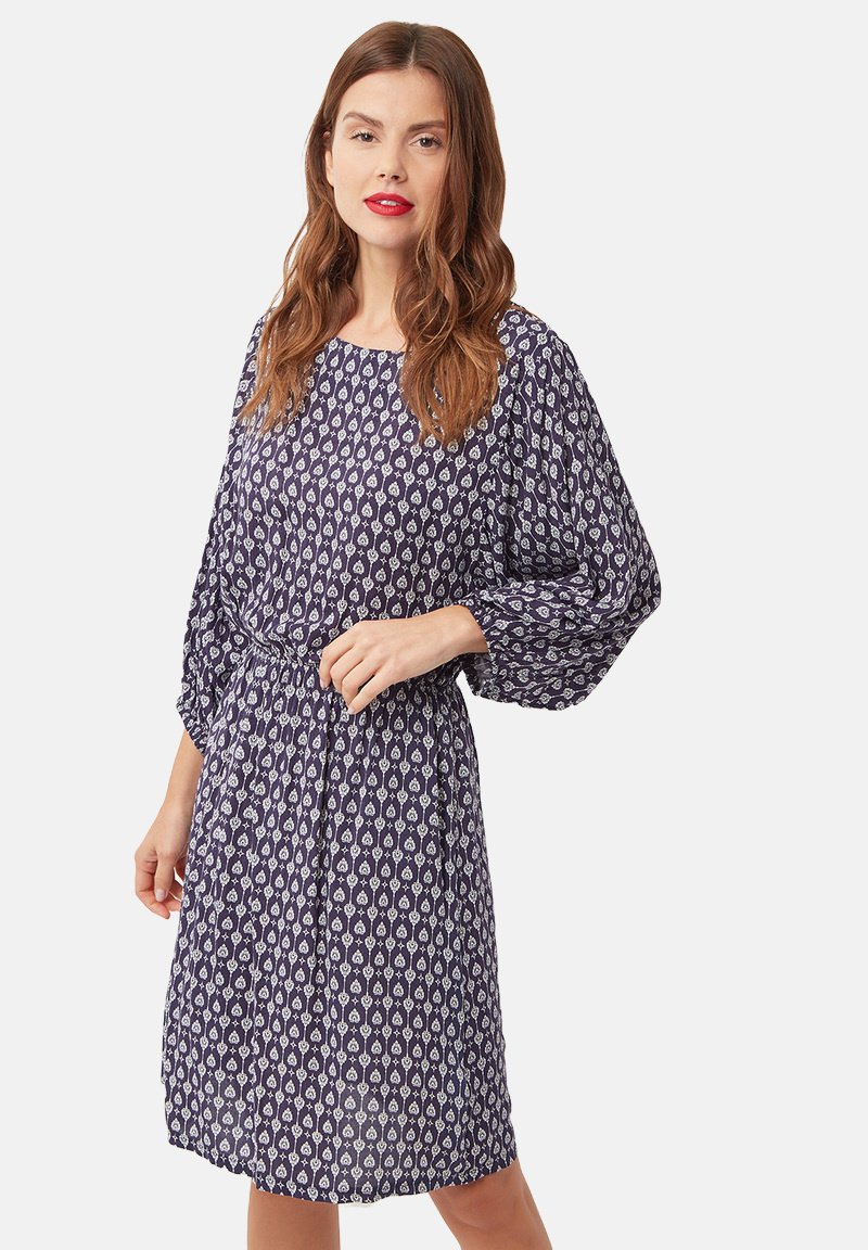 Mujer con cabello largo castaño que lleva un vestido azul marino con un estampado, de longitud hasta la rodilla y con mangas largas abullonadas, de pie frente a un fondo liso.