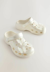 Weiße Kunststoffschuhe mit geschlossenem Zehenbereich und offenem Fersenbereich, ausgestattet mit Ausschnitten und drei schimmernden Schmetterlingsakzenten auf der Oberseite.