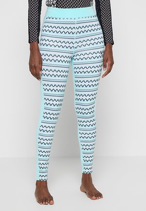 Leggings avec des panneaux latéraux bleu clair et un devant à motif zigzag en bleu marine et blanc, portés par une personne pieds nus debout sur un sol blanc.