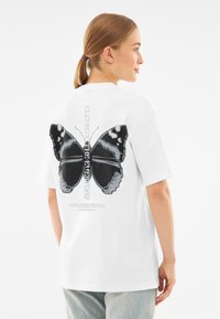 Camiseta blanca de manga corta con un gran gráfico de mariposa negra en la parte posterior, con texto debajo y un diseño de corte relajado.