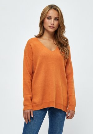 V-ringad orange stickad tröja med långa ärmar, avslappnad passform, ribbade muddar och nederkant. Matchad med blå jeans, mot en ljus bakgrund.