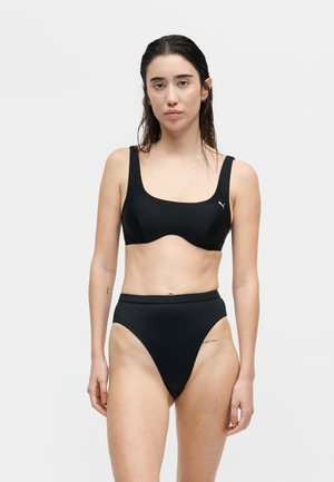 Γυναίκα με βρεγμένα μαλλιά, φορώντας μαύρο αθλητικό σουτιέν και μπικίνι με ψηλή μέση, στέκεται μπροστά σε ένα απλό λευκό φόντο.