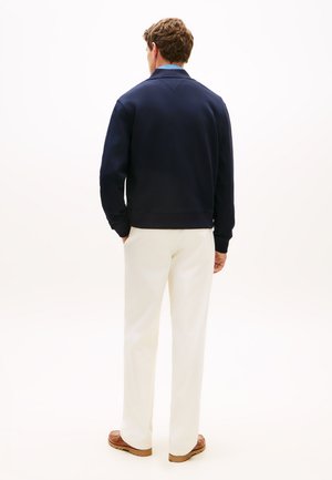 Muž otočený chrbtom, oblečený v námorníckej modrej bunde, bielych nohaviciach a hnedých topánkach na jednofarebnom svetlom pozadí.