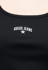 Μαύρο ριμπ ύφασμα τοπ με λευκό κείμενο "GUESS JEANS" και ένα αστέρι κεντημένο στο κέντρο κοντά στον λαιμό.