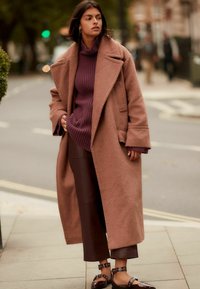 Abrigo marrón de gran tamaño en tejido texturizado, superpuesto a un suéter de punto morado grueso. La modelo lleva pantalones de cuero marrón oscuro y zapatos marrones brillantes.