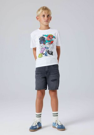 Ragazzo biondo in piedi con le mani dietro la schiena che indossa una maglietta bianca con grafica Fortnite, pantaloncini di jeans neri, calzini a righe e sneakers blu.