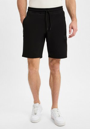 Pantalones cortos deportivos negros fabricados con tejido suave, con cintura elástica y cordón, que cuentan con bolsillos laterales y un parche de marca texturizado.