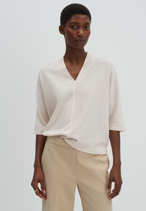 Femme portant un haut blanc ample à col en V avec des manches trois-quarts et un pantalon beige taille haute sur un fond gris clair.