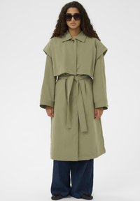 Olivno zeleni trench plašč s skrajšano zasnovo, širokim ovratnikom, pasom in dolgimi rokavi, v kombinaciji z širokimi jeans hlačami in črnimi čevlji.