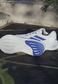White athletic shoes with blue accents, featuring a textured upper, cushioned white sole, and patterned blue outsole for traction.
