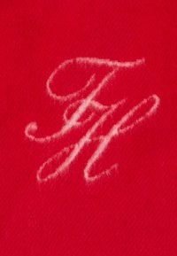 Raudonas audinys, turintis švelnų tekstūrą, su baltu išsiuvinėtu monogramu "FH" elegantišku kursyvu.