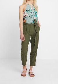 Top halter floral en azul-verde, con detalle de corte; pantalones de tiro alto en verde oliva con cinturón; sandalias rosa pálido con tiras.