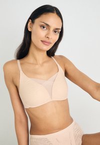 Beige myk stoff-BH med V-formet design, mesh-detalj, justerbare stropper og subtil tekstur med ribber. Glatt overflate, ingen synlige festemidler.