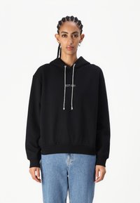 Svart hettegenser med snorer har et frontlogo "EST. 1981." Myk stoff, avslappet passform, kombinert med lyseblå jeans.