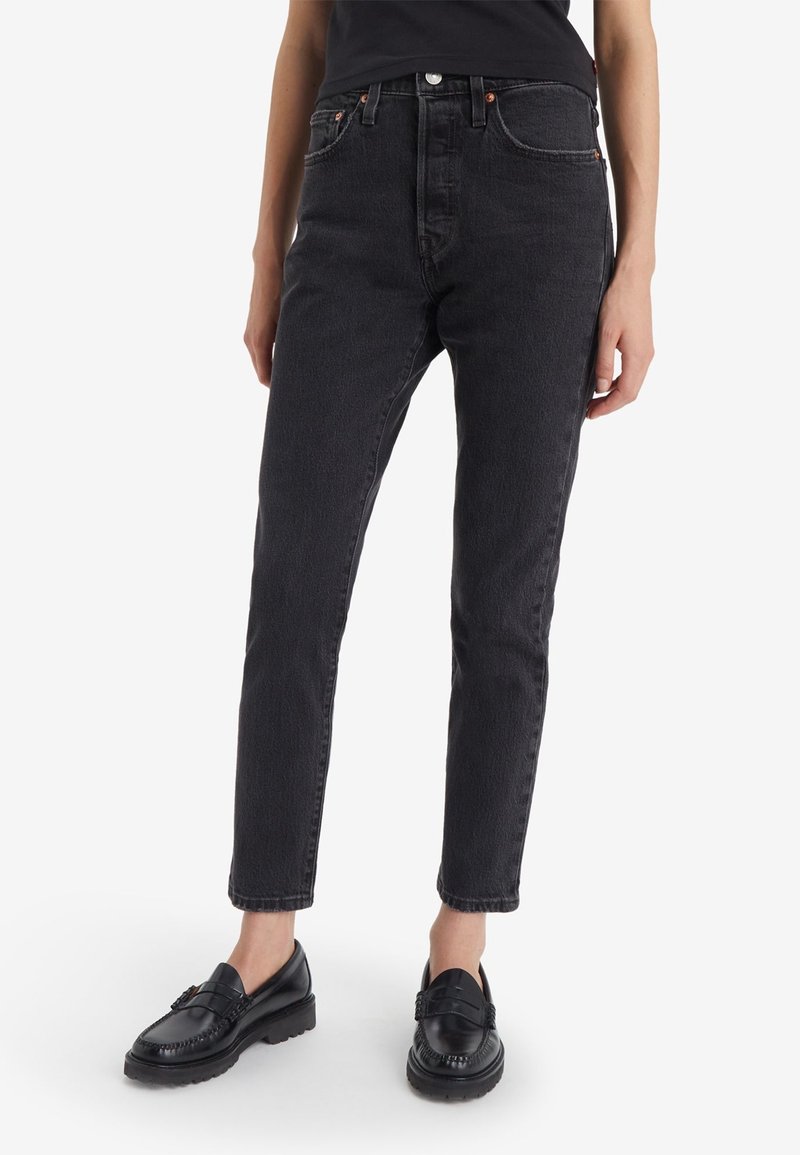 Personne portant un jean noir taille haute à jambes droites et des mocassins en cuir noir, debout devant un fond blanc.