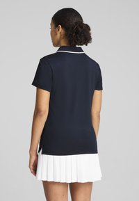 Dunkelblauses Polo-Shirt mit kurzen Ärmeln und Kragen sowie weißem Piping, kombiniert mit einem weißen, gefalteten Rock, der über das Knie reicht.