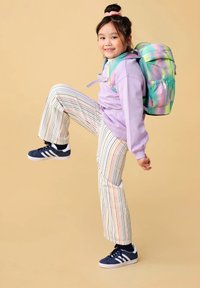 Jong meisje in gestreepte broek, lavendelkleurige hoodie en pastelrugzak tilt één been op en glimlacht tegen een beige achtergrond.