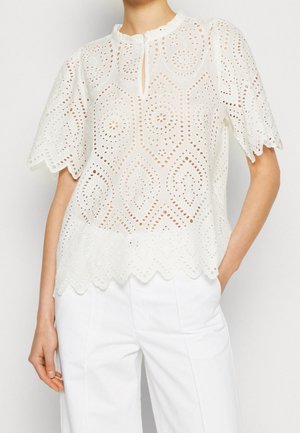 Woman wearing a white eyelet lace blouse with scalloped edges and short sleeves, paired with white high-waist trousers.