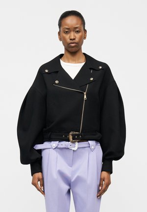 Γυναίκα που φοράει μαύρο φαρδύ πανωφόρι με φερμουάρ και φουσκωτά μανίκια, καθώς και ανοιχτό μωβ ψηλόμεσο παντελόνι με ζώνη.