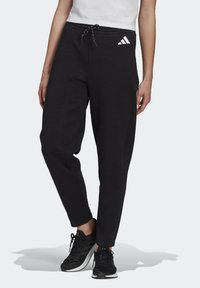 Persoon in zwarte Adidas trainingsbroek met een trekkoord in de taille en zwarte sportschoenen, staande tegen een effen achtergrond.