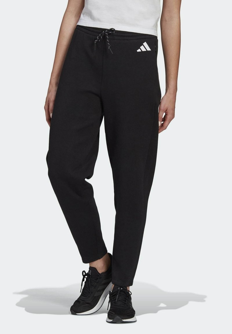 Persoon in zwarte Adidas trainingsbroek met een trekkoord in de taille en zwarte sportschoenen, staande tegen een effen achtergrond.