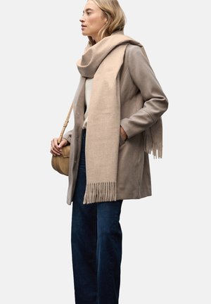 Beige uldfrakke med lommer, parret med et lysere beige tørklæde med frynse detaljer. Mørkeblå jeans og en lille brun taske fuldender looket.