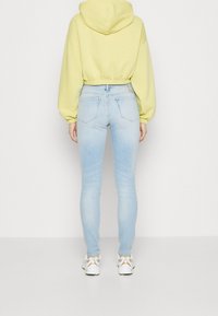 Sudadera recortada amarilla, jeans ajustados azul claro y zapatillas blancas con detalles en dorado. Tejido suave, puños elásticos y detalles en el bolsillo trasero visibles.