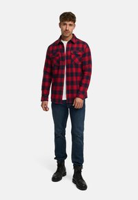 Chemise en flanelle à carreaux rouges et bleu marine avec deux poches poitrine, fermeture à boutons et manches longues, associée à un jean bleu foncé et des bottes noires.