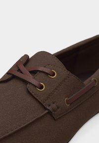 Καφέ σουέντ μοκασίνι με σκούρο καφέ δερμάτινη κορδέλα περασμένη μέσα από χρυσά μεταλλικά τρύπες, παρουσιάζεται σε κοντινή λήψη σε λευκό φόντο.