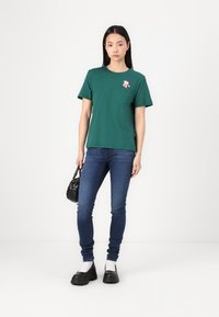 Grønn bomull t-skjorte med rund hals og lomme, prydet med et tegneserie-applikasjon, kombinert med blå smale jeans og svarte kraftige sko.