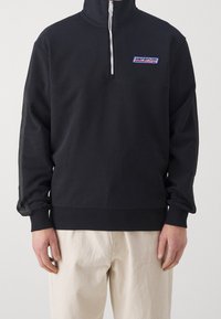 Sudadera negra con cremallera y cuello alto, que cuenta con puños y dobladillo acanalados, y un logotipo azul y blanco en el lado izquierdo del pecho. Tejido suave.